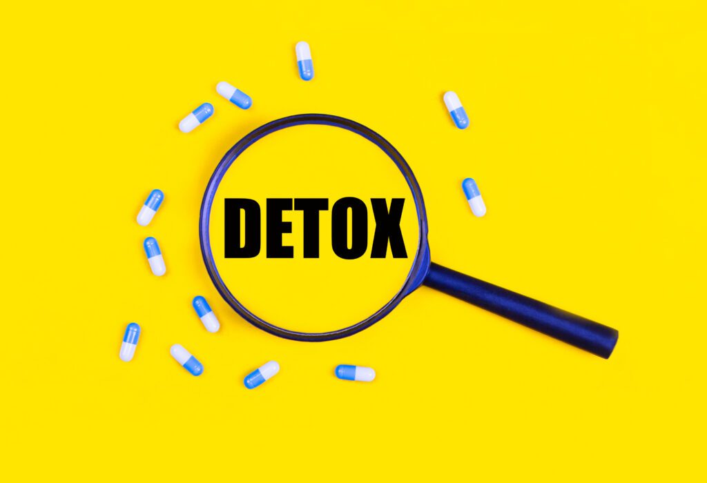 Pharmaceutical tablets and capsules near a magnifying glass with the text DETOX on a yellow background. View from above. Concepts of medicine.