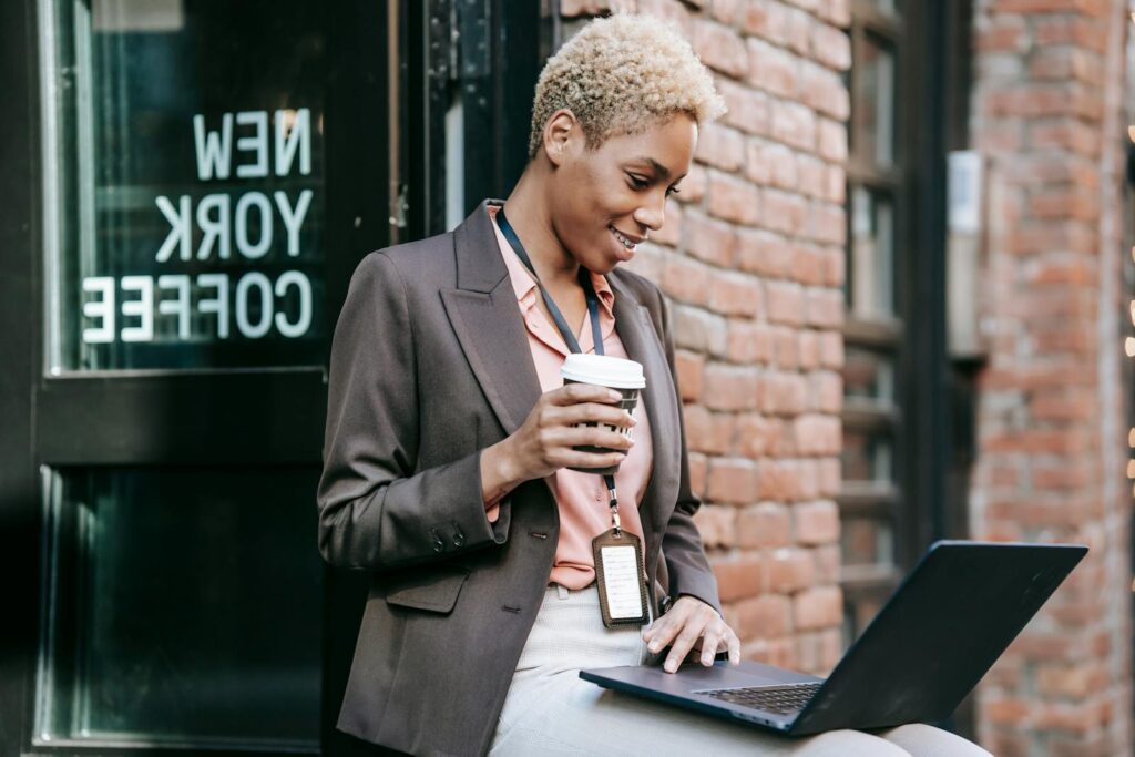 Positive African American businesswoman with cup of coffee browsing netbook in outdoor cafeteria while working remotely