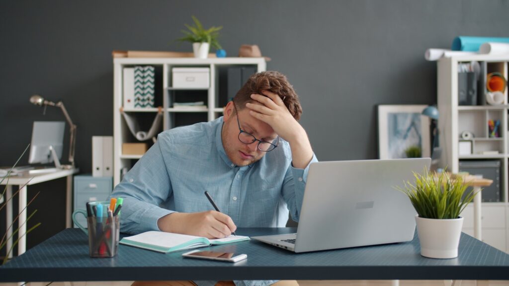 Dissatisfied With Your Job? Here’s a 5-Step Escape Plan 2 Man writing at desk with laptop, looking stressed.