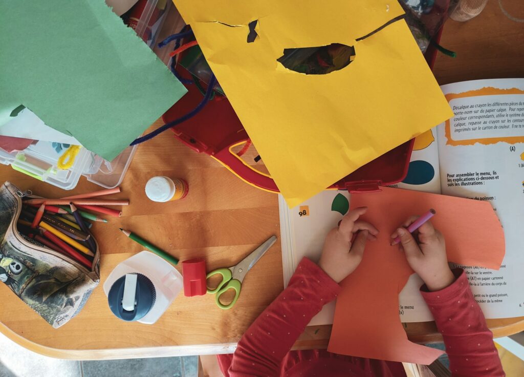 A child drawing on orange paper at a wooden table with craft supplies